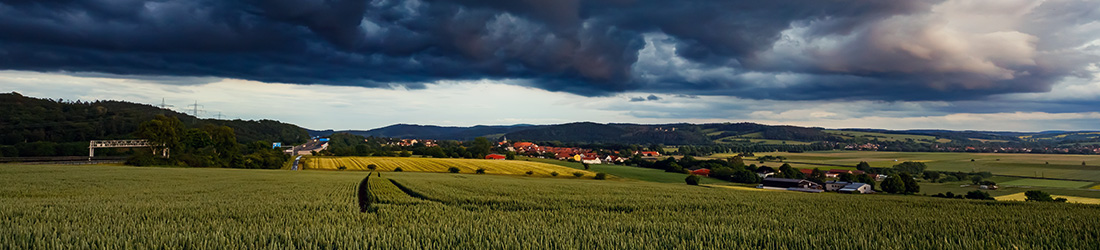 Eine sanft hügelige Landschaft mit grünen Feldern, im Hintergrund sehen wir höhere Berge. Darüber hängt eine bedrohliche Wolkendecke.