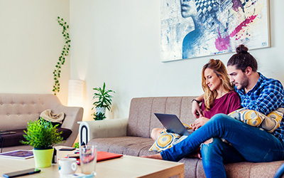 Ein junges Paar sitzt auf der Couch in einem modern eingerichten Wohnzimmer. Sie hat den Laptop auf dem Schoß und er hat den Arm um sie gelegt. Sie schließen vielleicht gerade die GEV Hausratversicherung online ab.