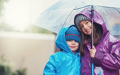 Zwei Kinder im Regen unter einem durchsichtigen Regenschirm. Das ältere – vermutliich ein Mädchen – hält den Schirm und trägt einen lila Regenmantel mit Kapuze. Das   kleinere – vermutlich ein Junge –  trägt ein einen blauen Regenmantel ebenfalls mit Kapuze. Beide haben passende Mützen unter den Kapuzen und blicken lächelnd in die Kamera.