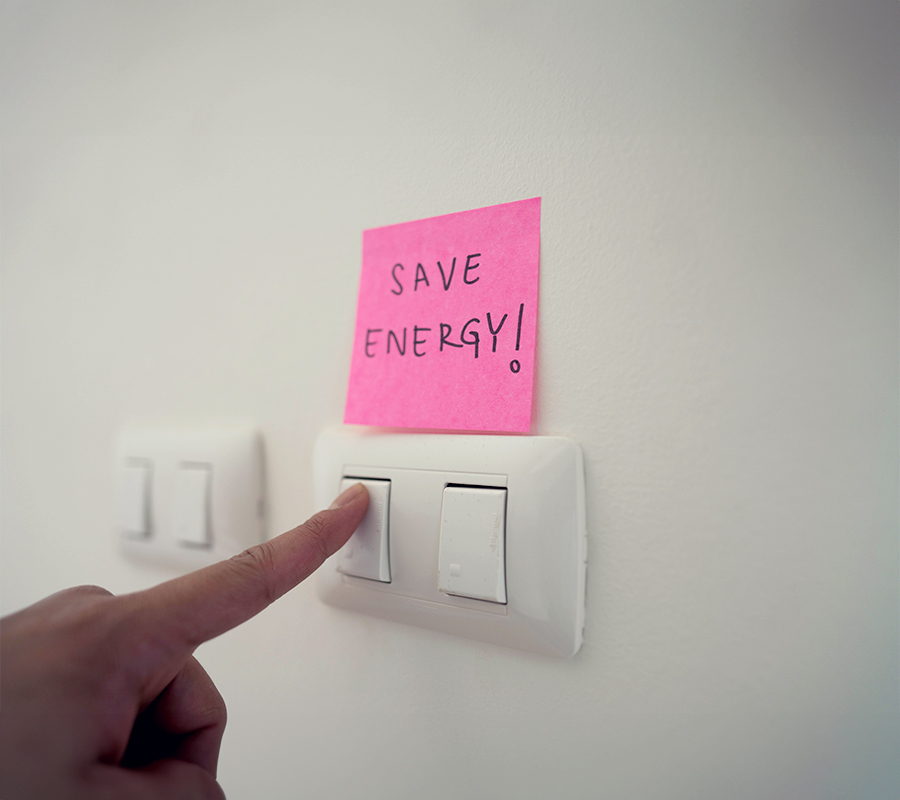 Zwei Doppellichtschalter. Über einem hängt ein Post-it mit der Aufschrift: Save Energy!. Ein Finger drückt auf einen Lichtschalter.