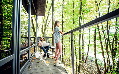 Eine Frau steht am Geländer eines großen Balkons. Das Haus ist modern und verfügt über große Glasflächen. Das Haus ist von großen Bäumen umgeben. Im Hintergrund sitzt ein junger Mann auf dem Balkon mit einem Laptop auf den Knien. 