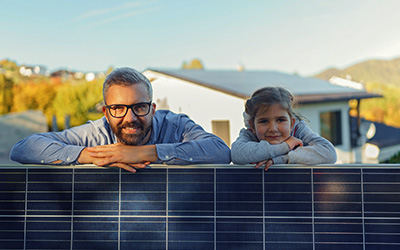 Vater und Tochter stehen hinter einem Solarpanel und stützen sich darauf ab. Im Hintergrund sehen wir ihr Haus. 