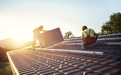 Zwei Fachleute verlegen Solarpanels auf einem Dach im Gegenlicht der untergehenden Sonne.