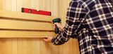 Sauna selbst bauen leicht gemacht – und danach entspannt ins Schwitzen kommen Person beim Bau einer Heimsauna: Holzverkleidung wird mit einer Bohrmaschine an der Wand befestigt und mithilfe einer Wasserwaage exakt ausgerichtet.
