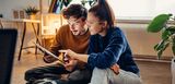 Ein junger Mann und eine junge Frau schauen sich gemeinsam auf dem Boden sitzend etwas auf einem Tablet an.